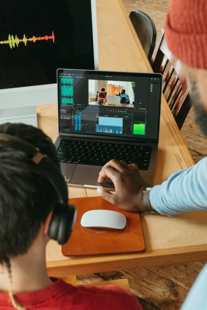 Two People Working On Video Editing At A Wooden Desk. One Is Wearing Headphones, The Other Is Using A Laptop With A Video Timeline Displayed While Researching &Quot;What Is Closed Captioning.&Quot; A Larger Monitor Shows Audio Waveforms.