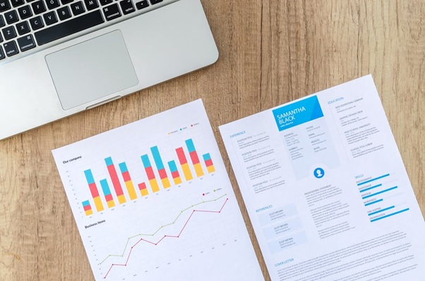 A Laptop On A Wooden Desk Sits Beside Two Documents: A Colorful Bar And Line Graph Chart Illustrating Online Marketing Trends, And A Resume Highlighting Experience, Education, And Skills.